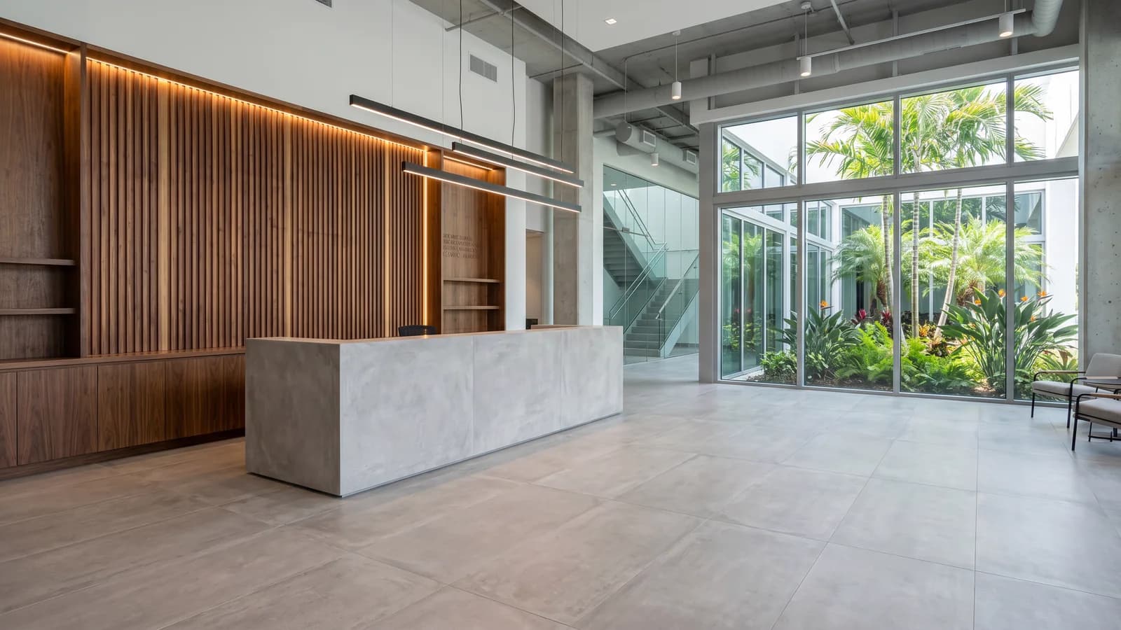 Lobby corporativo en Aventura con piso de porcelanato look concreto matte gris claro de gran formato, escritorio de recepción revestido en mismo porcelanato, pared de nogal, iluminación negra matte, vista a patio con palmeras